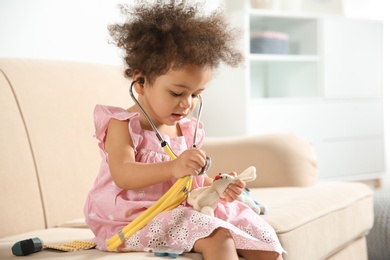 Photo of Cute African American child imagining herself as doctor while playing with stethoscope and toy on couch at home
