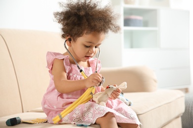 Cute African American child imagining herself as doctor while playing with stethoscope and toy on couch at home