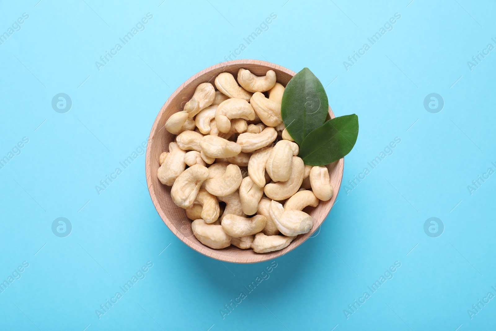 Photo of Tasty cashew nuts and green leaves in bowl on light blue background, top view