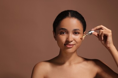 Beautiful woman applying serum onto her face on brown background. Space for text
