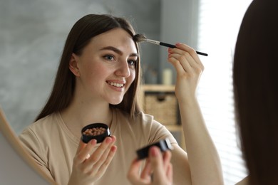 Photo of Smiling woman with freckles applying makeup near mirror indoors