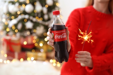 Photo of MYKOLAIV, UKRAINE - January 01, 2021: Woman with bottle of Coca-Cola and sparkler against blurred Christmas tree, closeup