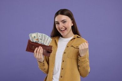 Excited woman holding wallet with dollar banknotes on purple background