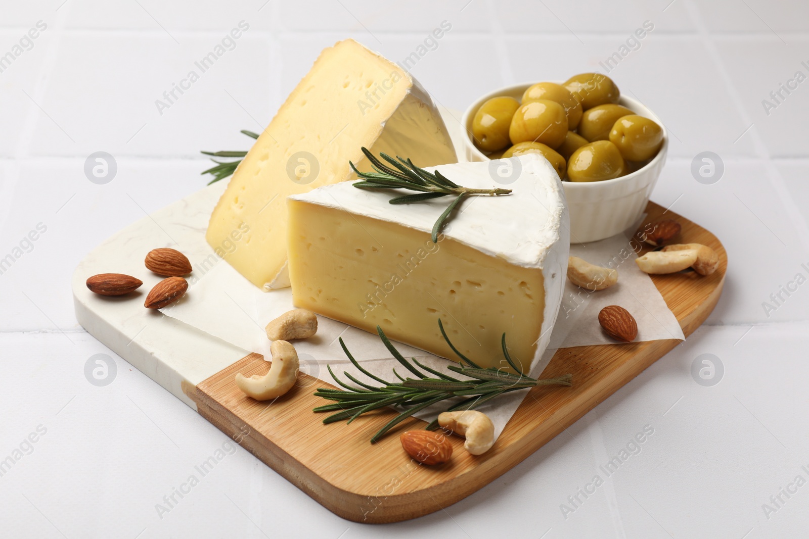 Photo of Pieces of tasty camembert cheese, rosemary, nuts and olives on white tiled table, closeup