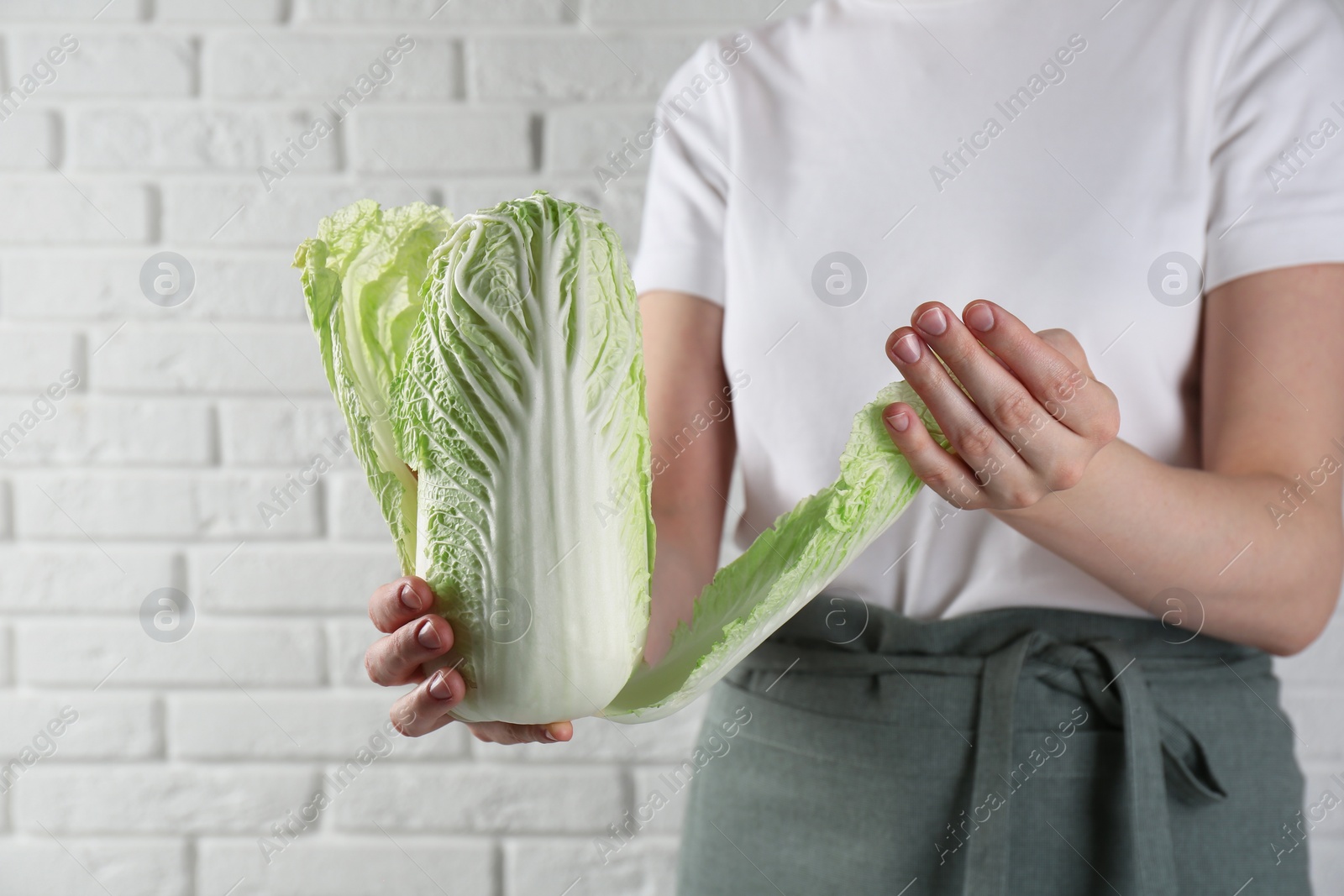 Photo of Woman separating leaf from fresh Chinese cabbage near white brick wall, closeup