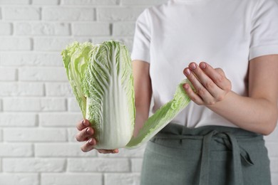 Photo of Woman separating leaf from fresh Chinese cabbage near white brick wall, closeup