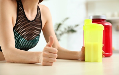 Young woman with bottle of protein shake at table in kitchen, closeup view
