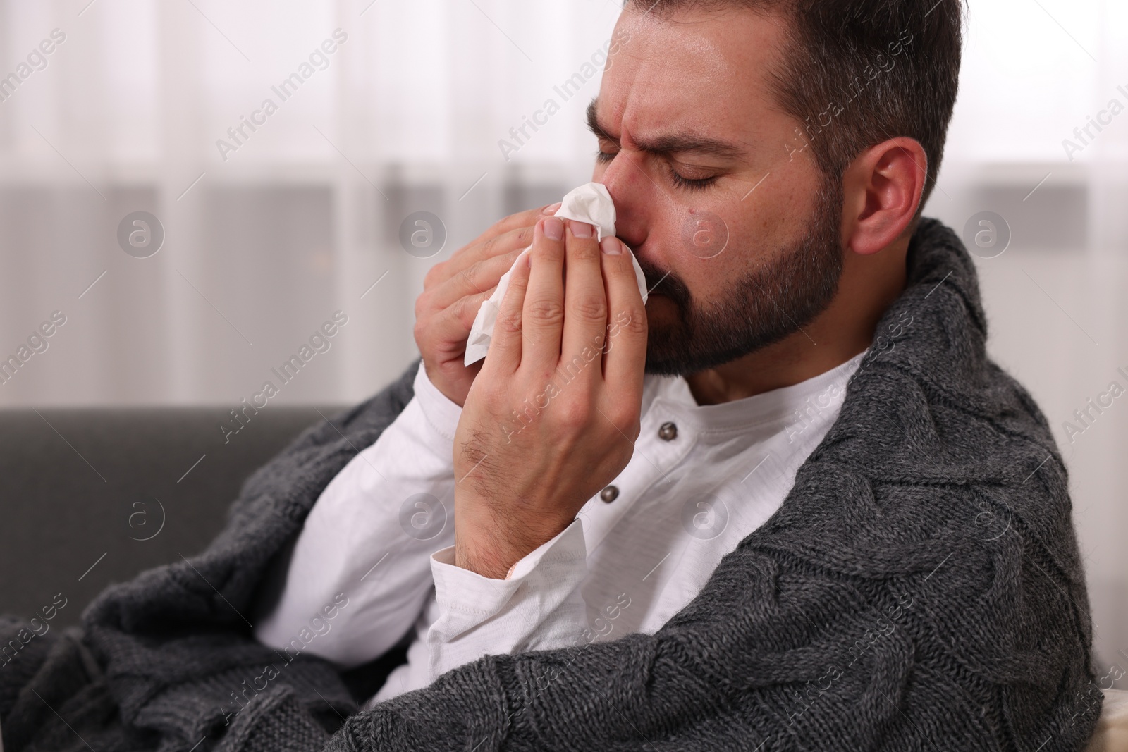 Photo of Sick man wrapped in blanket with tissue blowing nose on sofa indoors. Cold symptoms