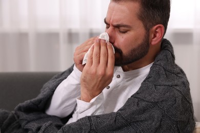 Photo of Sick man wrapped in blanket with tissue blowing nose on sofa indoors. Cold symptoms