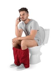 Young man with mobile phone sitting on toilet bowl. Isolated on white