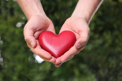 Young man holding red heart on blurred green background, closeup. Donation concept