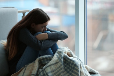 Photo of Upset teenage girl sitting at window indoors. Space for text