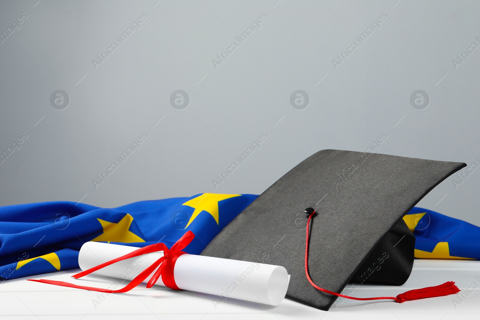 Photo of Black graduation cap, diploma and flag of European Union on white wooden table against grey wall