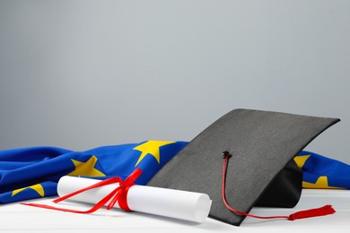 Photo of Black graduation cap, diploma and flag of European Union on white wooden table against grey wall