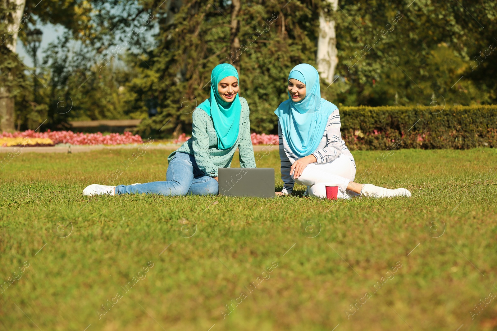 Photo of Muslim women in hijabs with laptop sitting on green lawn outdoors