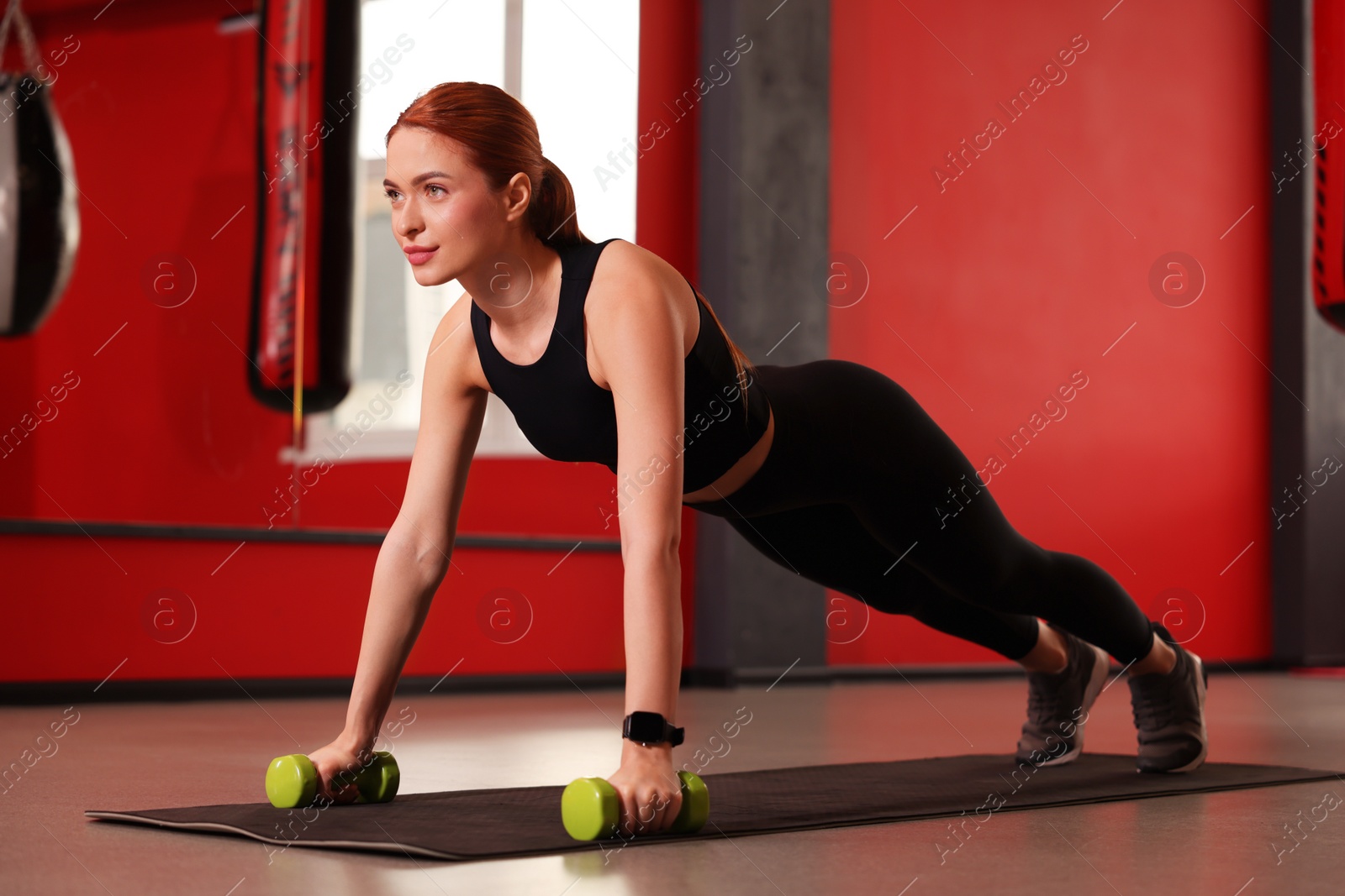 Photo of Athletic young woman with dumbbells doing plank exercise on mat in gym