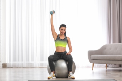 Photo of Young woman doing exercise with dumbbell on fitness ball at home