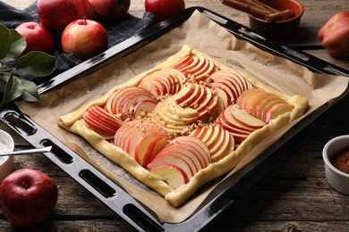 Photo of Baking tray with uncooked apple galette and ingredients on wooden table, closeup