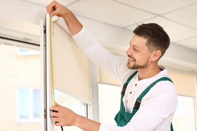 Handyman with tape measure installing roller window blind indoors