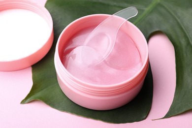 Photo of Under eye patches in jar with spatula and green leaf on light pink background, closeup. Cosmetic product