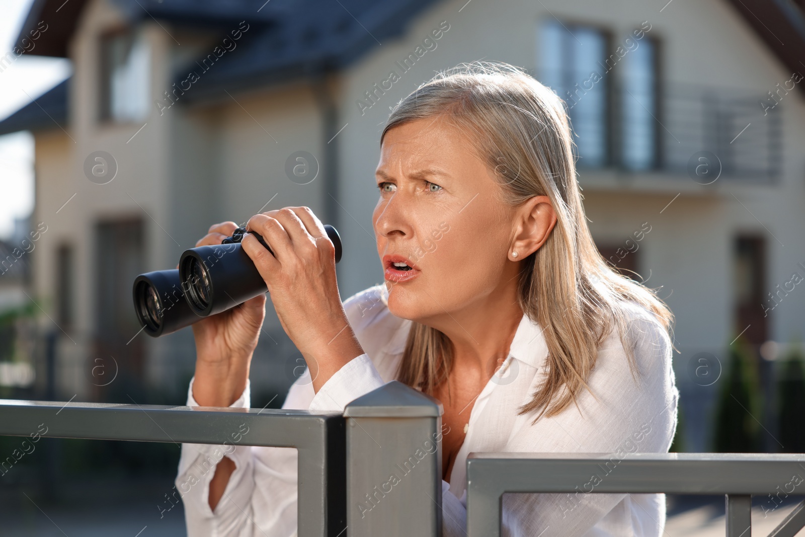 Photo of Concept of private life. Curious senior woman with binoculars spying on neighbours over fence outdoors