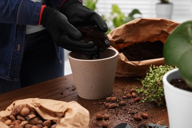 Photo of Woman in gloves filling flowerpot with soil at wooden table indoors, closeup. Transplanting houseplants