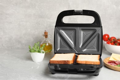 Modern sandwich maker with bread slices and different products on light grey table, space for text