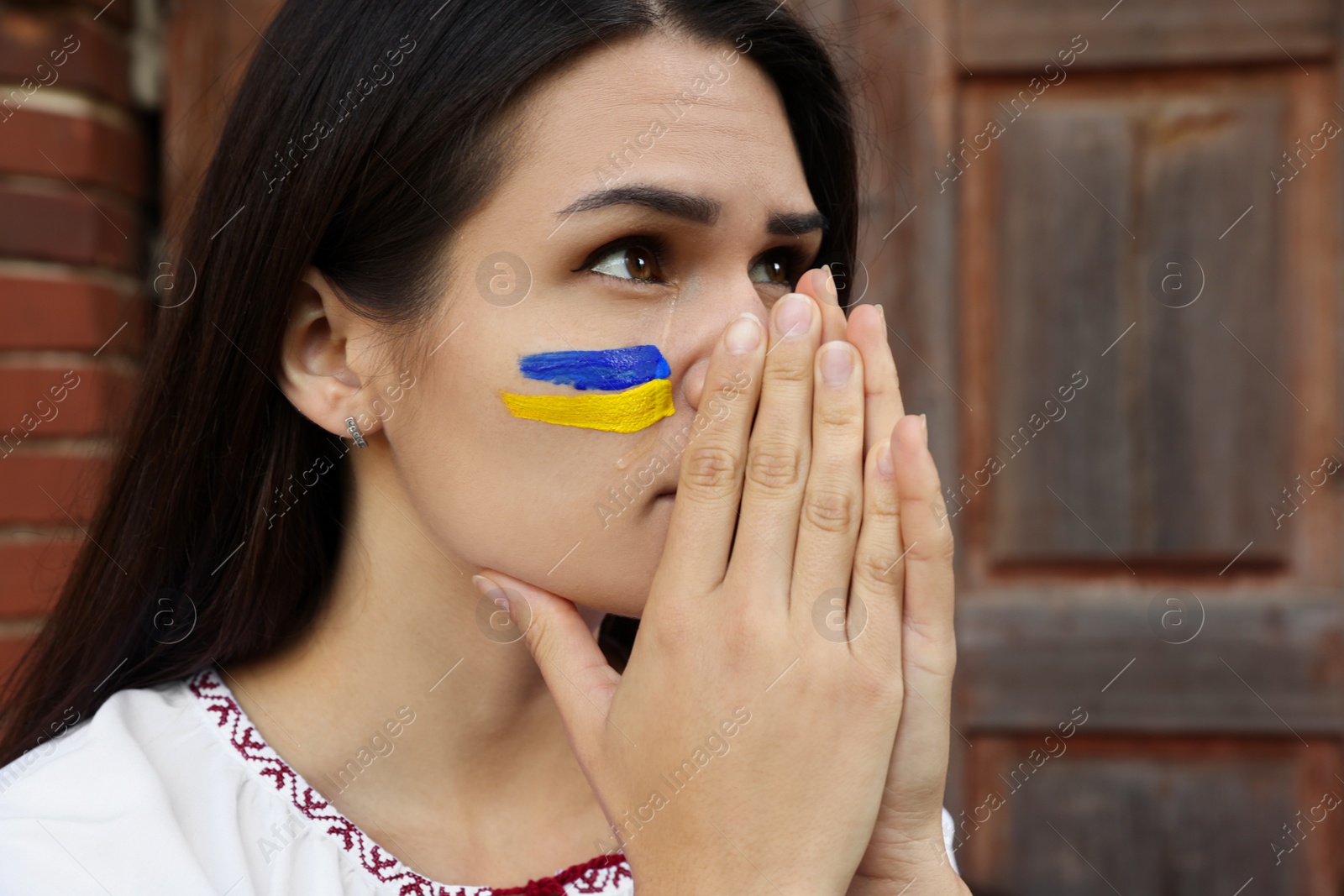 Photo of Sad young woman with drawing of Ukrainian flag on face near wooden door, closeup