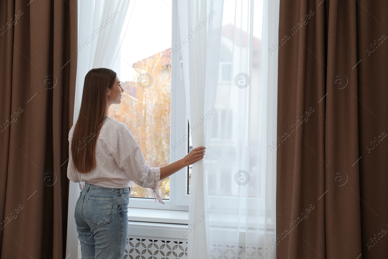 Photo of Woman opening elegant window curtains in room, back view