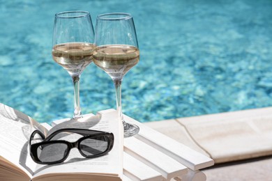 Photo of Glasses of tasty wine, sunglasses and open book on swimming pool edge. Space for text
