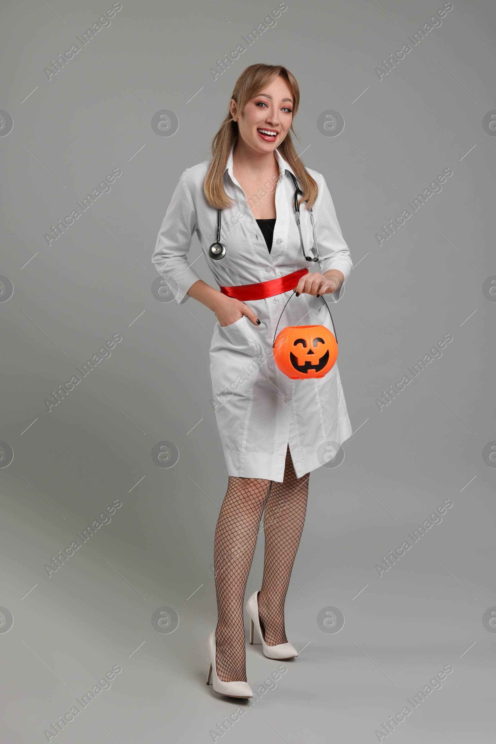 Photo of Happy woman in scary nurse costume with pumpkin bucket on light grey background. Halloween celebration