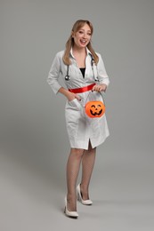 Happy woman in scary nurse costume with pumpkin bucket on light grey background. Halloween celebration