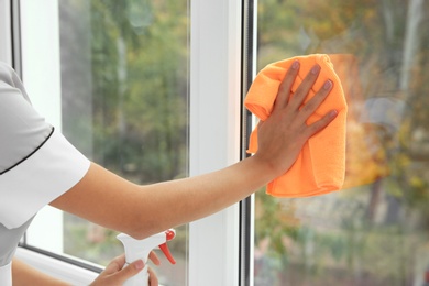 Photo of Young chambermaid cleaning window with rag indoors, closeup