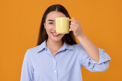 Happy young woman covering eye with yellow ceramic mug on orange background