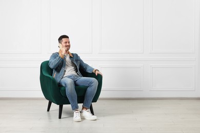 Happy man sitting in armchair and talking on smartphone indoors, space for text