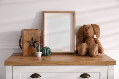 Photo of Empty photo frame near cute toy bunny and decor on dresser, space for text. Baby room interior element