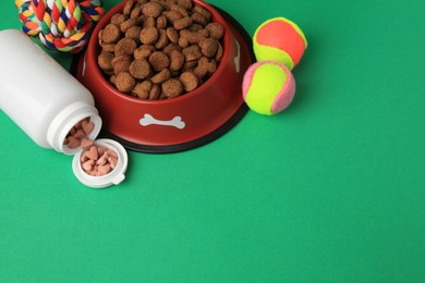 Photo of Bowl with dry pet food, bottle of vitamins and toys on green background. Space for text