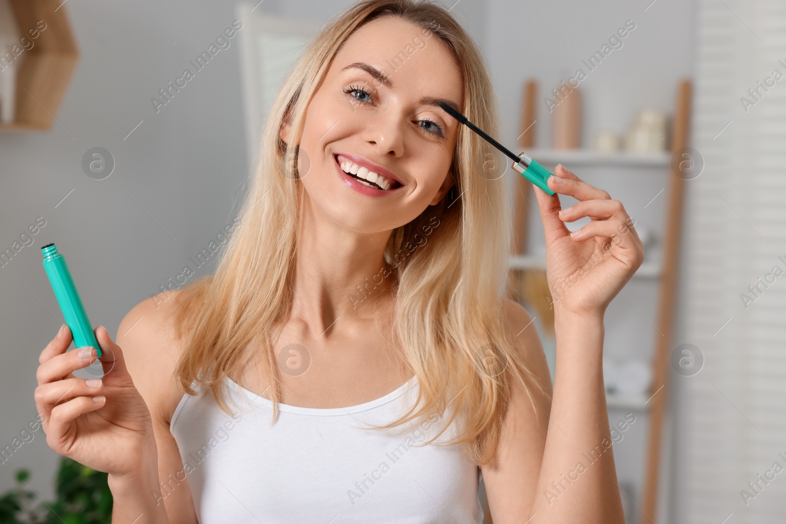 Photo of Beautiful happy woman applying mascara in bathroom