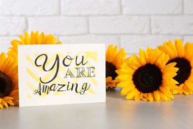 Photo of Card with life-affirming phrase You Are Amazing and sunflowers on light table against white brick wall
