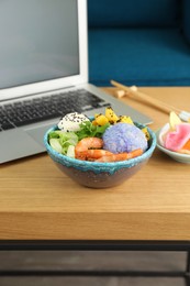 Delicious poke bowl near laptop on wooden table