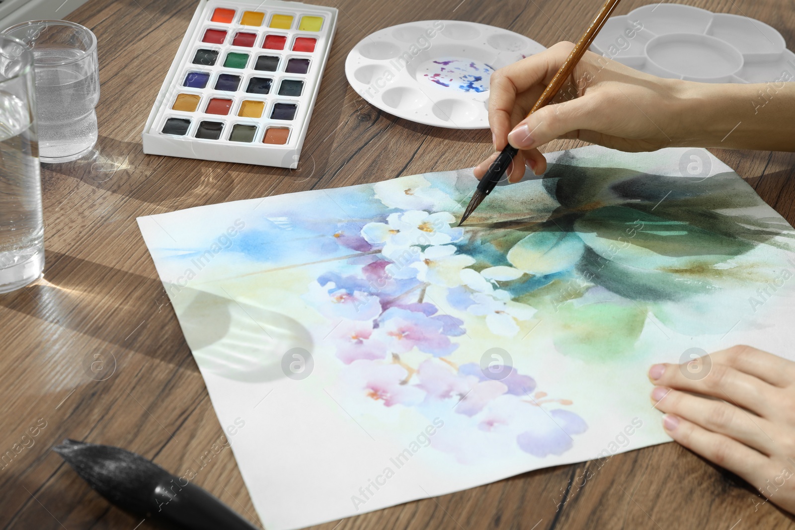 Photo of Woman painting flowers with watercolor at wooden table, closeup. Creative artwork