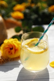 Composition with glass of refreshing lemonade and beautiful rose on white wooden table in blooming garden