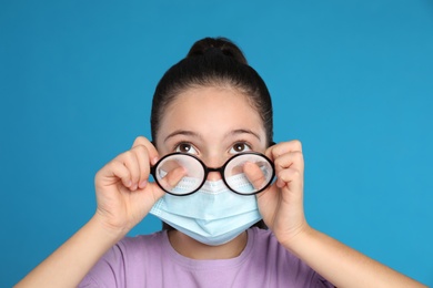 Photo of Little girl wiping foggy glasses caused by wearing medical face mask on blue background. Protective measure during coronavirus pandemic