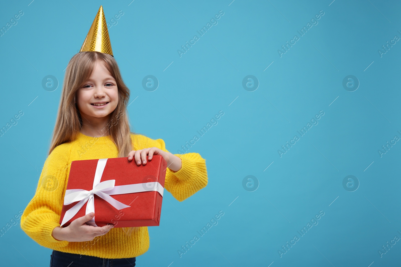 Photo of Happy little girl in party hat with gift on light blue background. Space for text