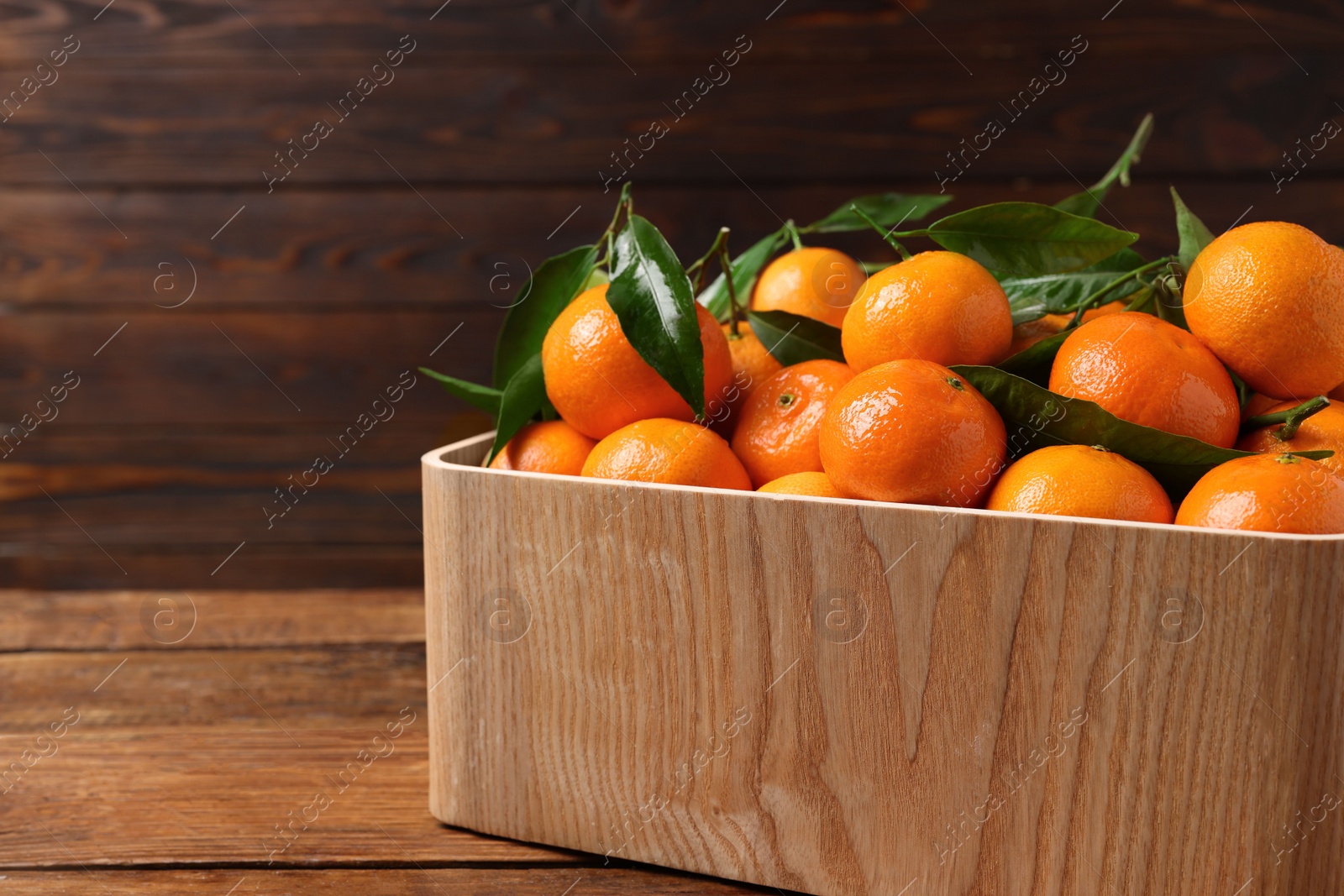 Photo of Fresh tangerines with green leaves in crate on wooden table, closeup. Space for text