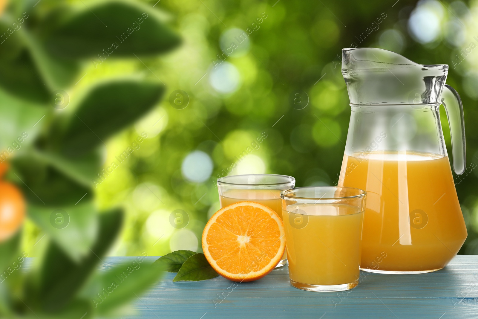 Image of Fresh orange juice on light blue wooden table in orchard. Space for text