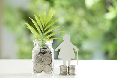 Photo of Pension savings. Figure of senior woman, jar with coins and twig on white table against blurred green background