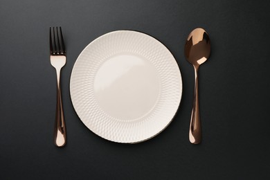 Photo of Empty plate, golden fork and spoon on black table, top view