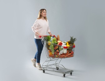 Photo of Young woman with shopping cart full of groceries on grey background