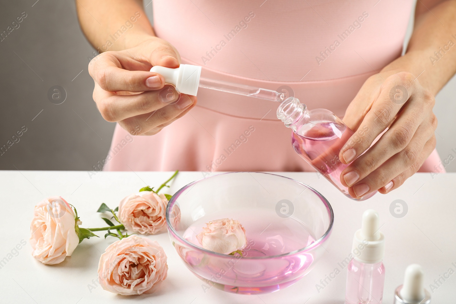 Photo of Young woman dripping rose essential oil into glass bottle at table, closeup